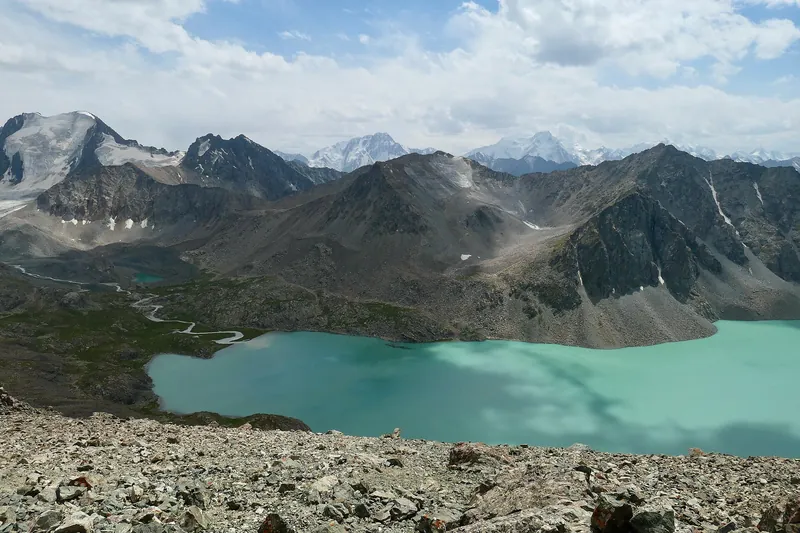 Draufsicht auf einen türkisfarbenen See zwischen felsigen Bergen, über den Gipfeln schweben Wolken.