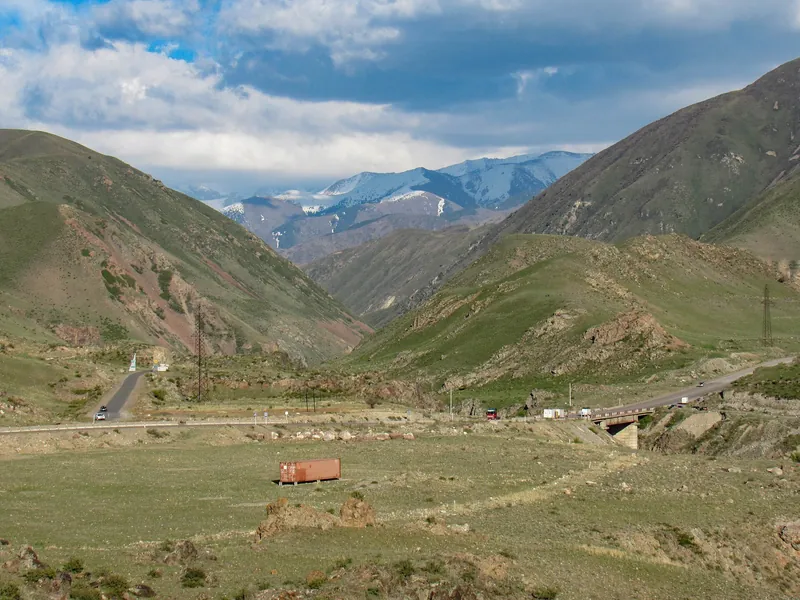 La strada attraversa una verde valle di montagna, con cime innevate visibili in lontananza dietro i dolci pendii.