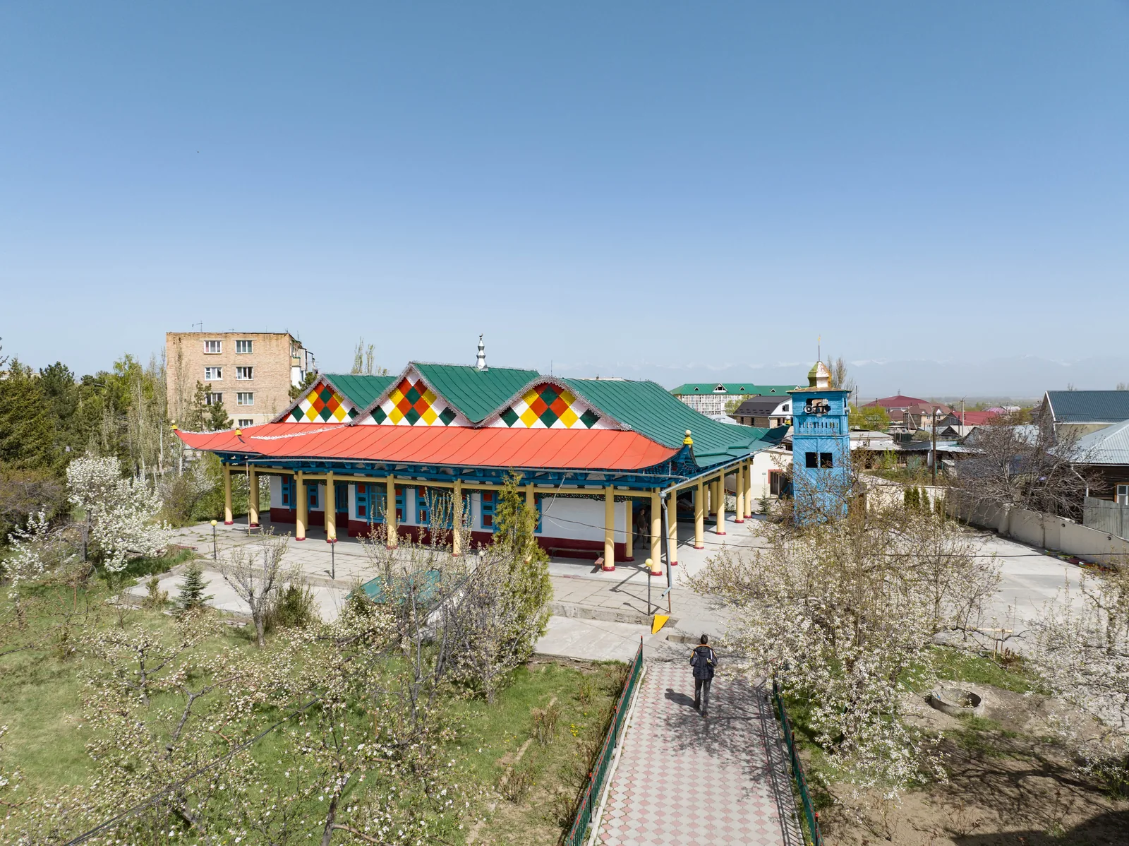 Moschee mit bunten Giebeln und blauem Minarett hinter einem blühenden Garten; ein Weg führt dorthin