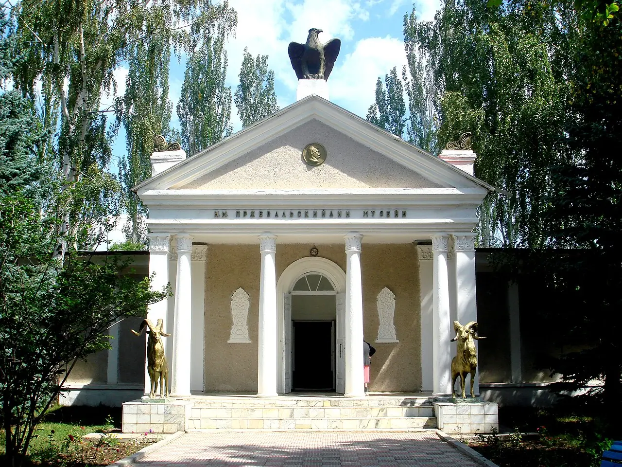 Pórtico blanco con columnas y un águila de bronce sobre el frontón, rodeado de árboles
