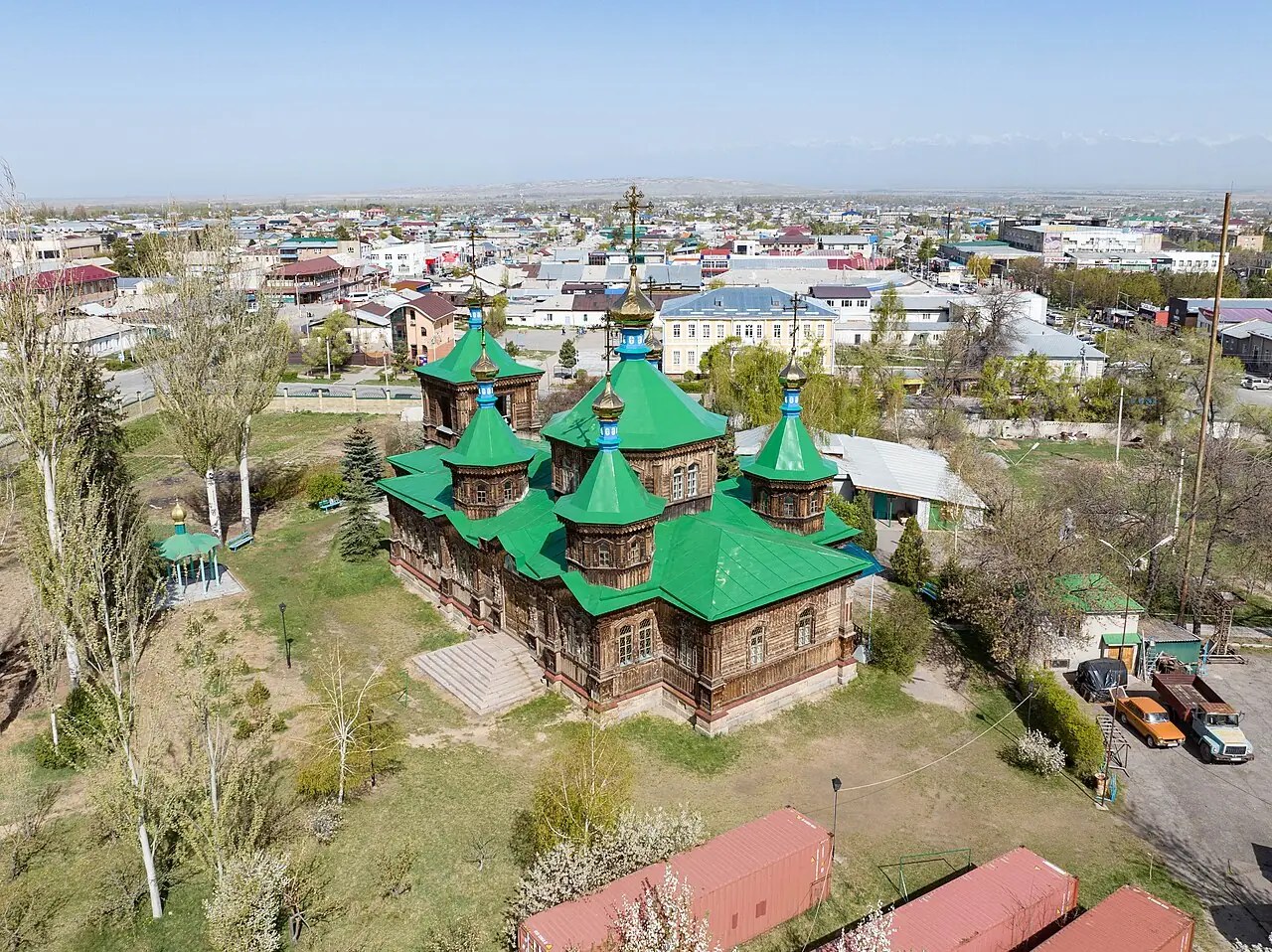 Desde arriba: catedral de cúpulas verdes entre árboles en flor, con tejados y montañas detrás