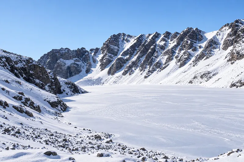 Verschneite Oberfläche eines Bergsees unter felsigen Bergrücken und klarem Winterhimmel.