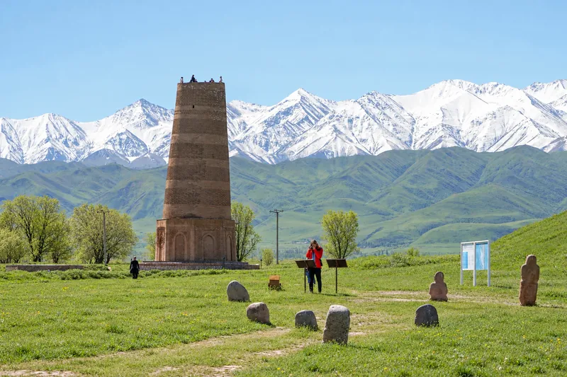 Brick tower and stone sculptures on a green lawn in front of a snowy mountain range; spring view.