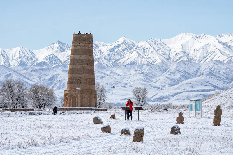 Brick tower and stone sculptures on a green lawn in front of a snowy mountain range; winter view.