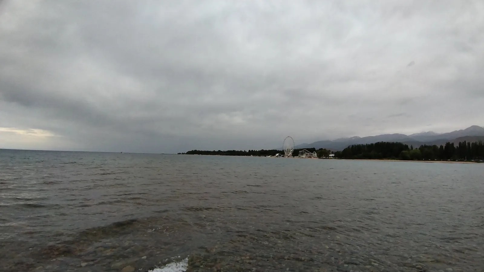 See am flachen Ufer unter grauen Wolken, in der Ferne ist ein Gebirgszug kaum zu erkennen.