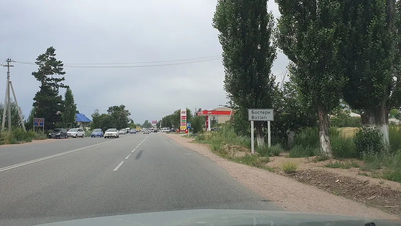 Eine breite Landstraße führt unter bewölktem Himmel zwischen Bäumen und niedrigen Gebäuden hindurch.