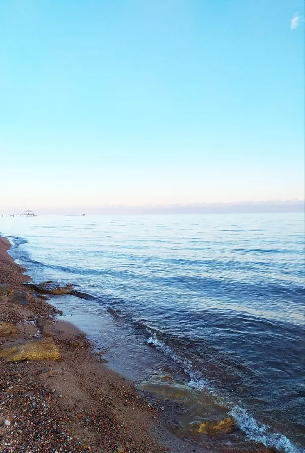 Kleine Wellen schlagen auf das felsige Ufer, ein heller Streifen Himmel liegt über dem blauen Wasser.