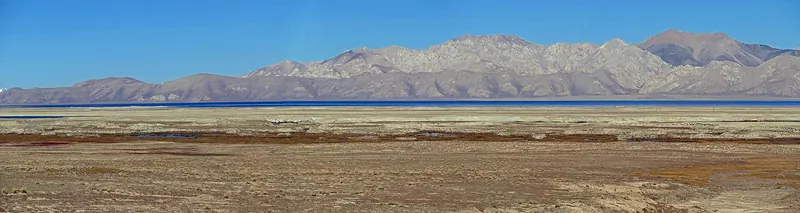 Una striscia di lago d’alta quota oltre una riva piatta e bruna, con montagne chiare in lontananza.
