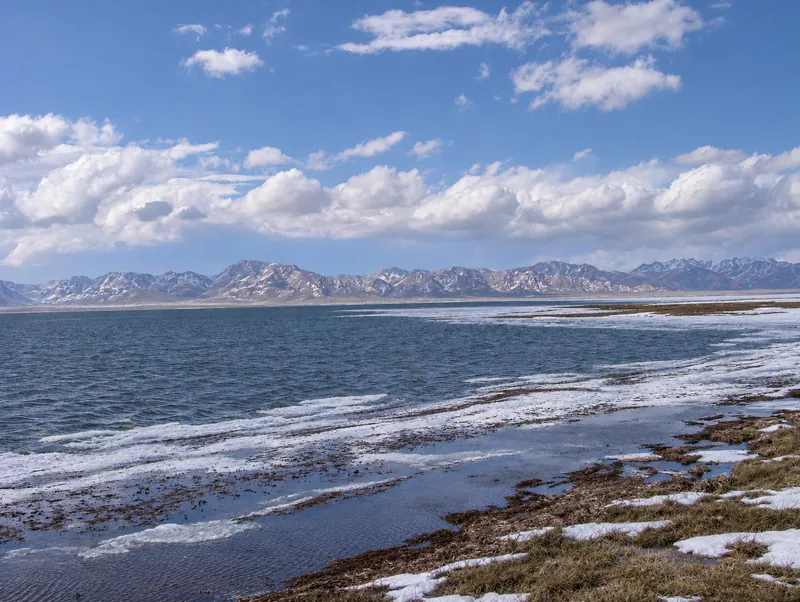 Riva primaverile del lago Chatyr-Kol con resti di neve e una catena montuosa dietro l'acqua.