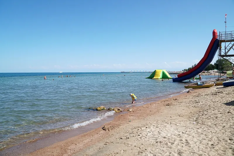 Eine rote Wasserrutsche steht am Sandufer mit Booten und flachem, klarem Seewasser.