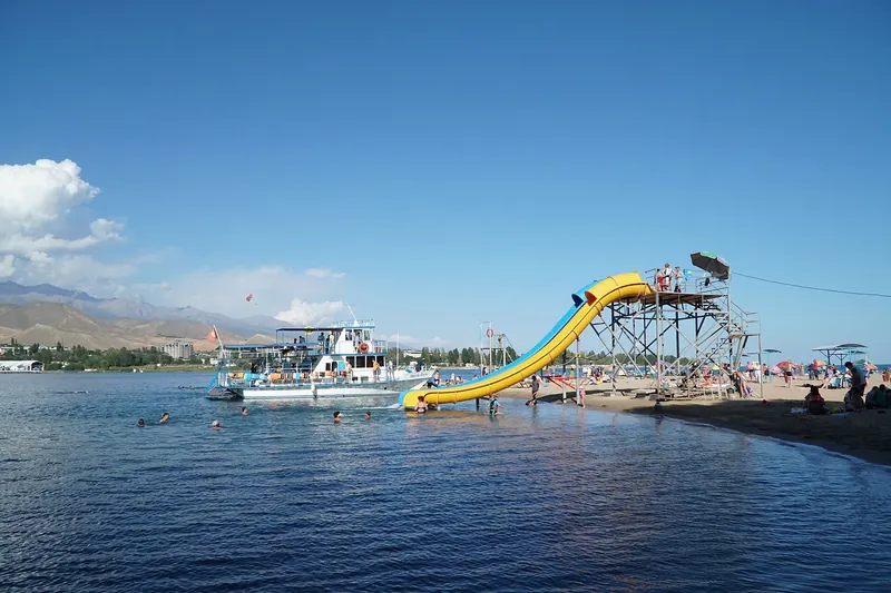 Eine gelbe Wasserrutsche führt vom Pier zum Wasser; Hinter den Booten ist ein fernes Bergufer sichtbar.