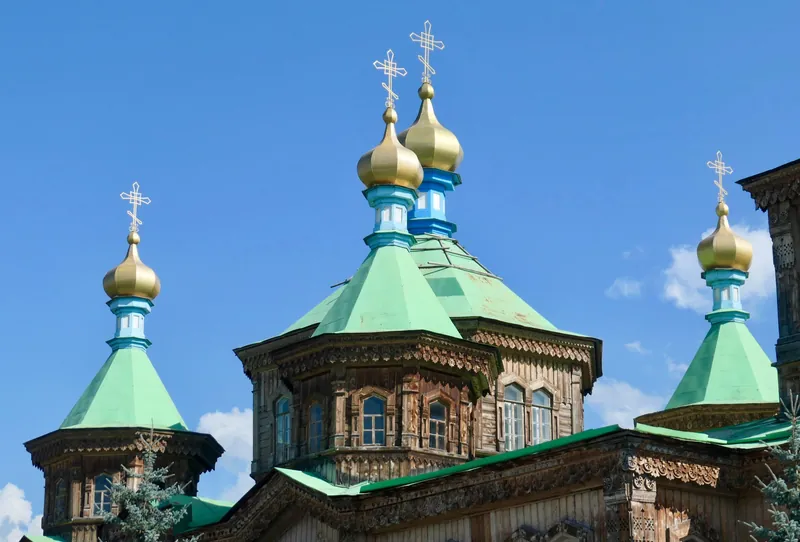 Goldene Kreuze und Kuppeln einer Holzkirche über grünen Dächern vor dem Himmel.