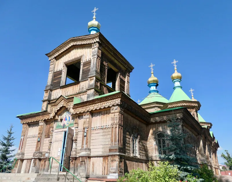 Ecke eines hölzernen Glockenturms mit geschnitzter Fassade, grünem Dach und goldener Kuppel.