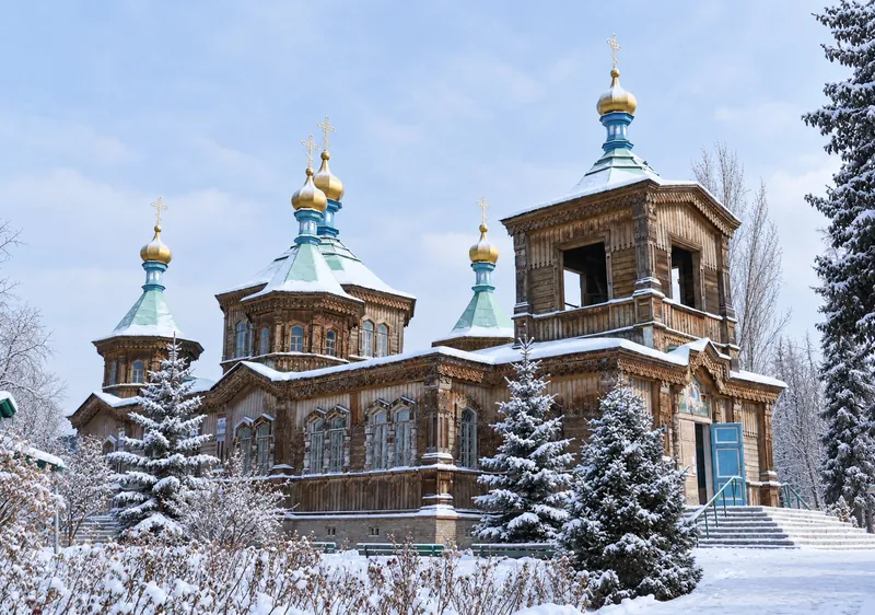 Holzkirche mit grünen Dächern und goldenen Kuppeln hinter einem Blumengarten; Winteransicht.