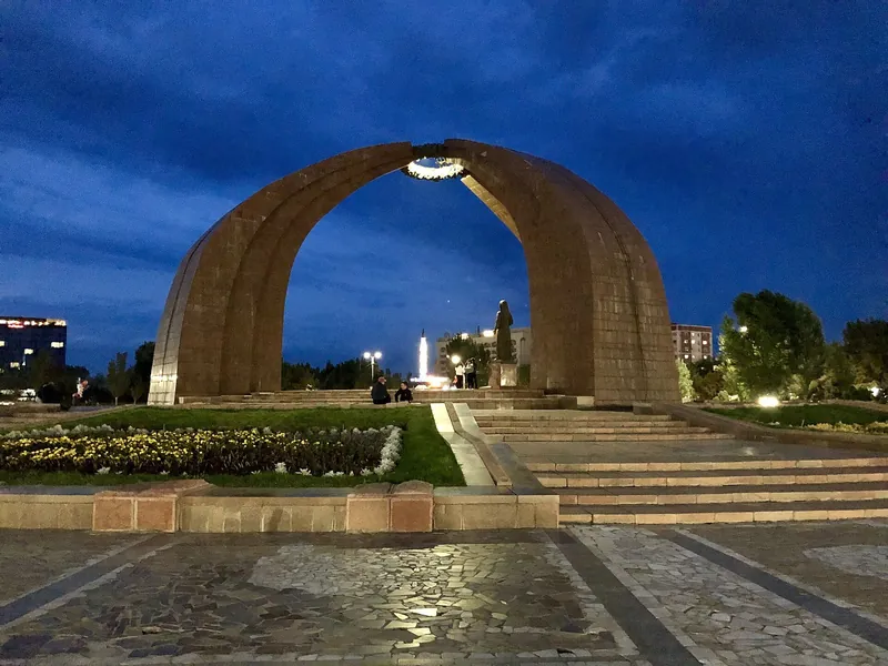 Bögen des Denkmals am Siegesplatz im blauen Abendlicht, davor Stufen und Blumenbeete.