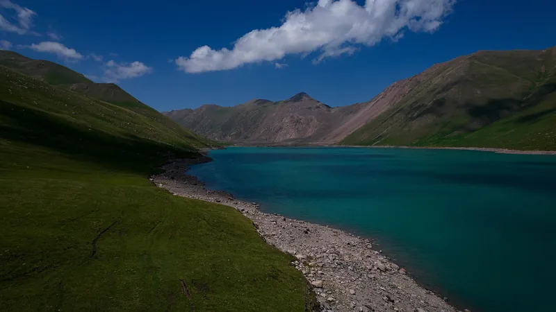 Blaues Wasser am flachen Seeufer, dahinter grüne Berghänge.