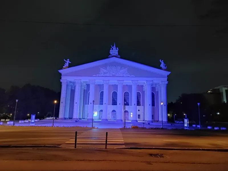 Die Fassade des Opern- und Balletttheaters in der abendlichen violetten Beleuchtung hinter einer breiten Stadtstraße.