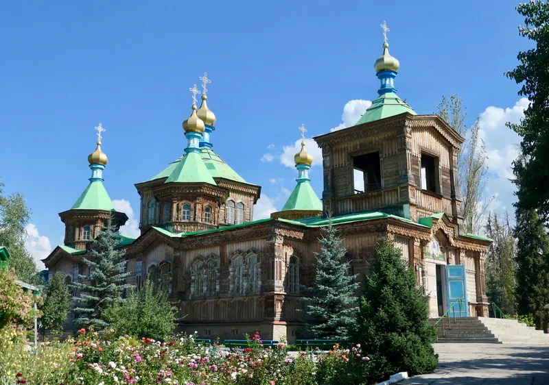 Iglesia de madera con tejados verdes y cúpulas doradas detrás de un jardín de flores.
