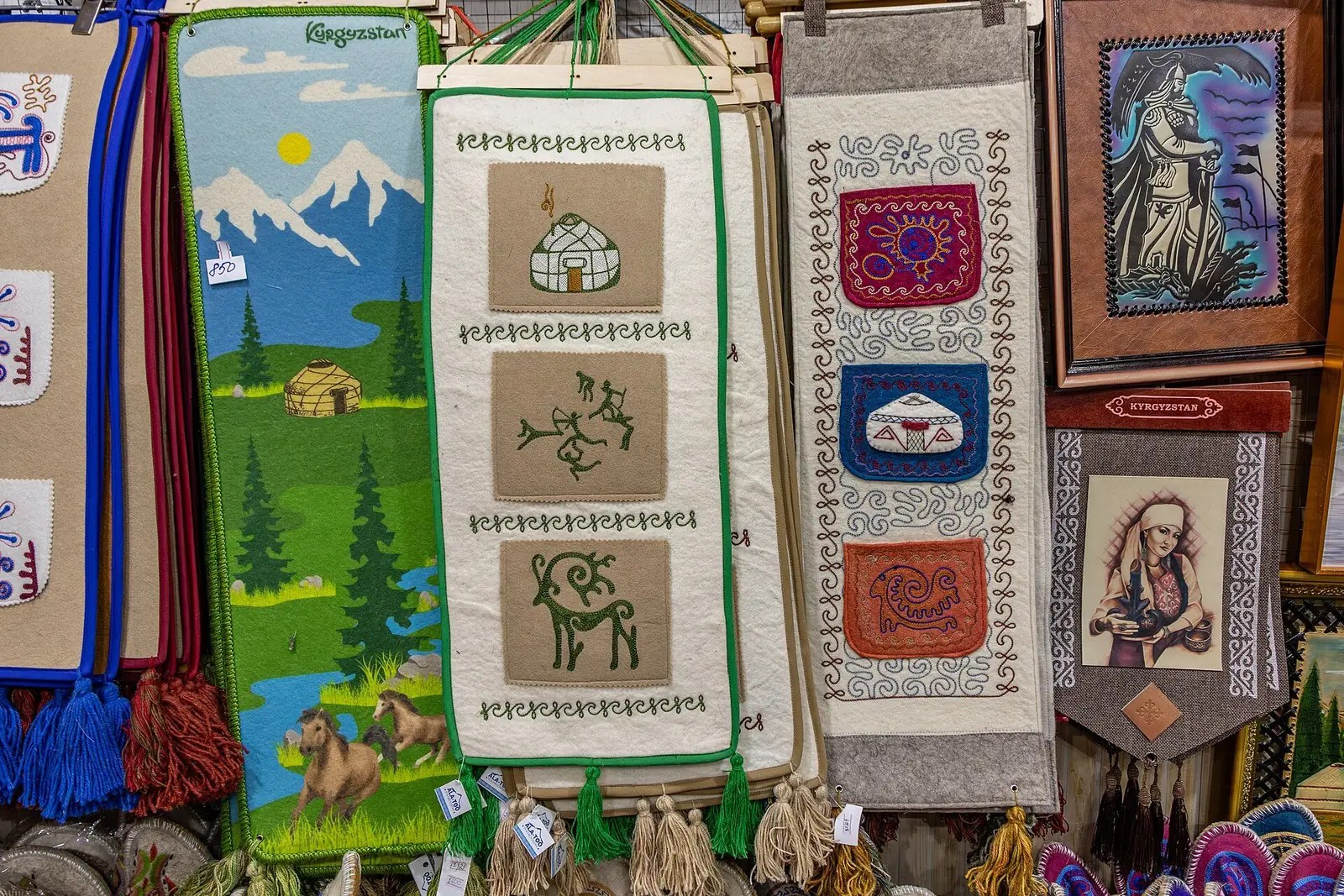 Bunte Filzbilder mit Ornamenten und szenischen Darstellungen hängen in einer Marktstandreihe.
