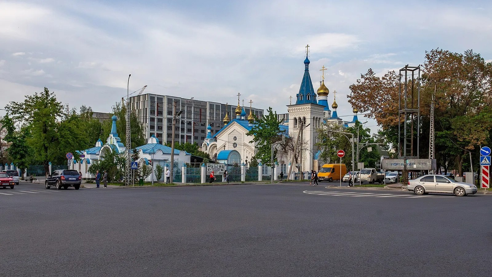 Eine weiße Kirche mit blauen Kuppeln hinter Zaun und Bäumen; davor eine breite Stadtstraße.