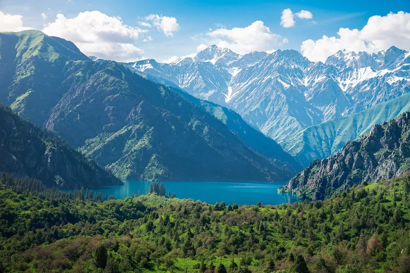 Türkisfarbener See zwischen Waldhängen, dahinter ein langer verschneiter Kamm.