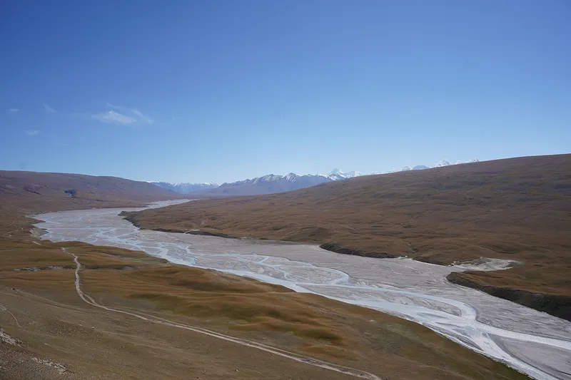 Ein grauer Fluss verzweigt sich im breiten Tal, hinter trockenen Hängen sind Schneeberge zu sehen.