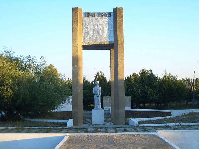 Памятник под высокой прямоугольной аркой, вокруг деревья и открытая площадка.