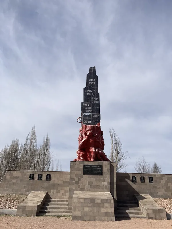 Eine schwarze Stele mit Datteln erhebt sich über roten Figuren und Steinstufen, umgeben von kahlen Bäumen.