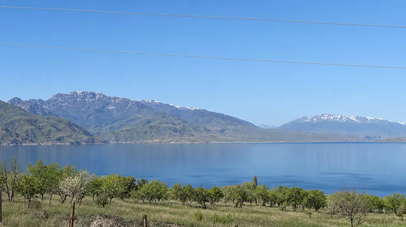 Toktogul-Stausee im Frühling, grüne Gärten am Ufer und Bergketten jenseits des Wassers.