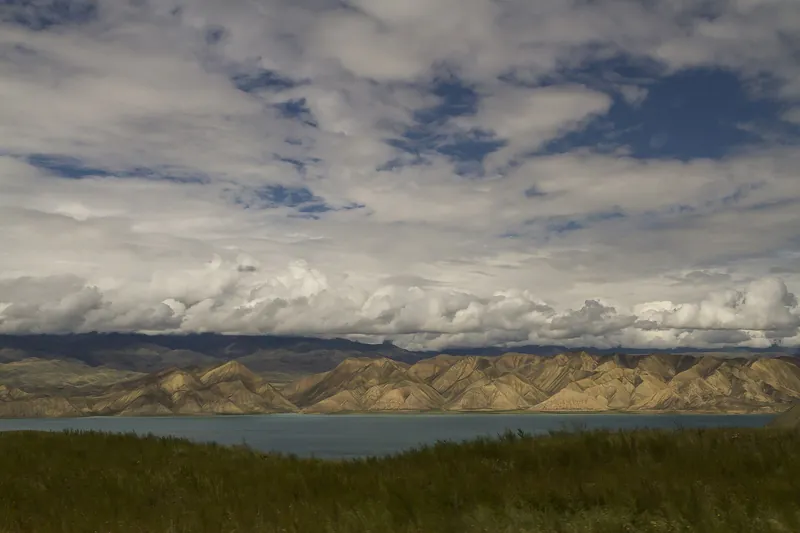Dunkle Wolken hängen über den hellen Berghängen in der Nähe des Stausees hinter der grünen Küstenwiese.