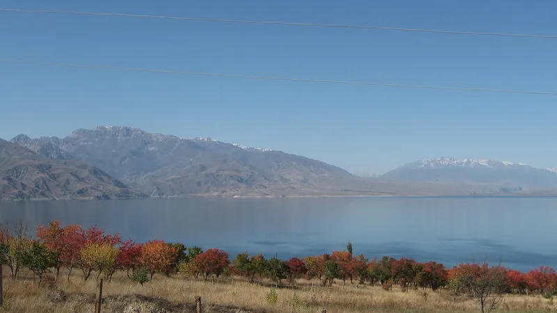 Herbstbäume über dem ruhigen Wasser des Stausees, hinter dem gegenüberliegenden Ufer Berge.