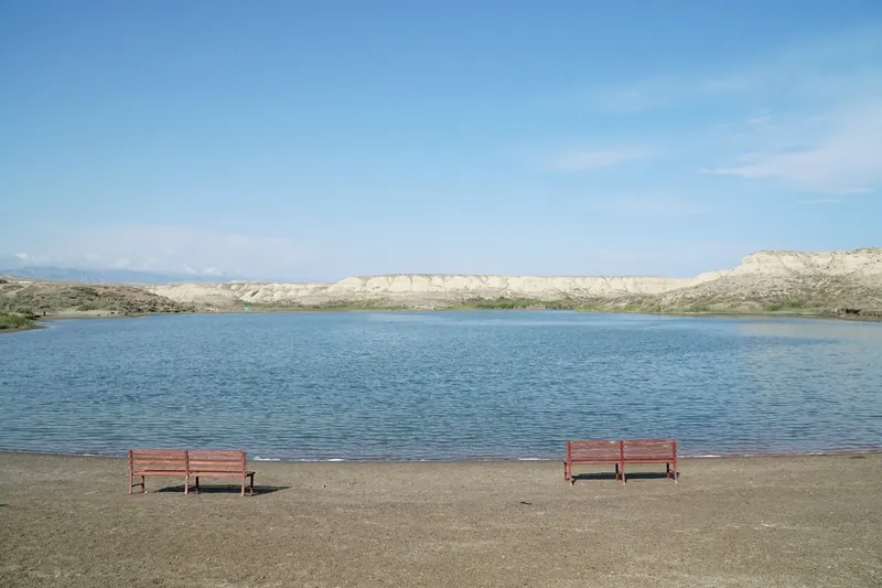 Две красные скамейки на ровном берегу Туз-Кёля перед голубой водой и белыми холмами.