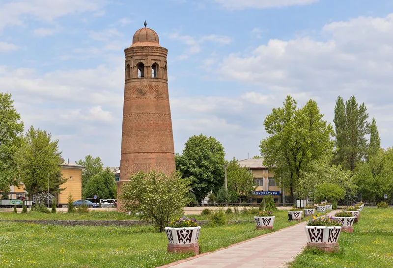 Un minaret de briques à maçonnerie ornée et sommet arrondi au-dessus de la pelouse et des massifs; vue au printemps.