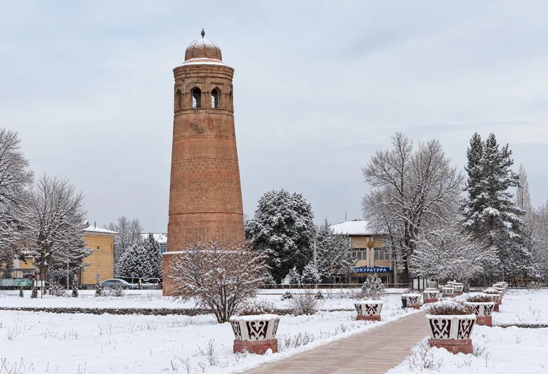 Un minaret de briques à maçonnerie ornée et sommet arrondi au-dessus de la pelouse et des massifs; vue en hiver.