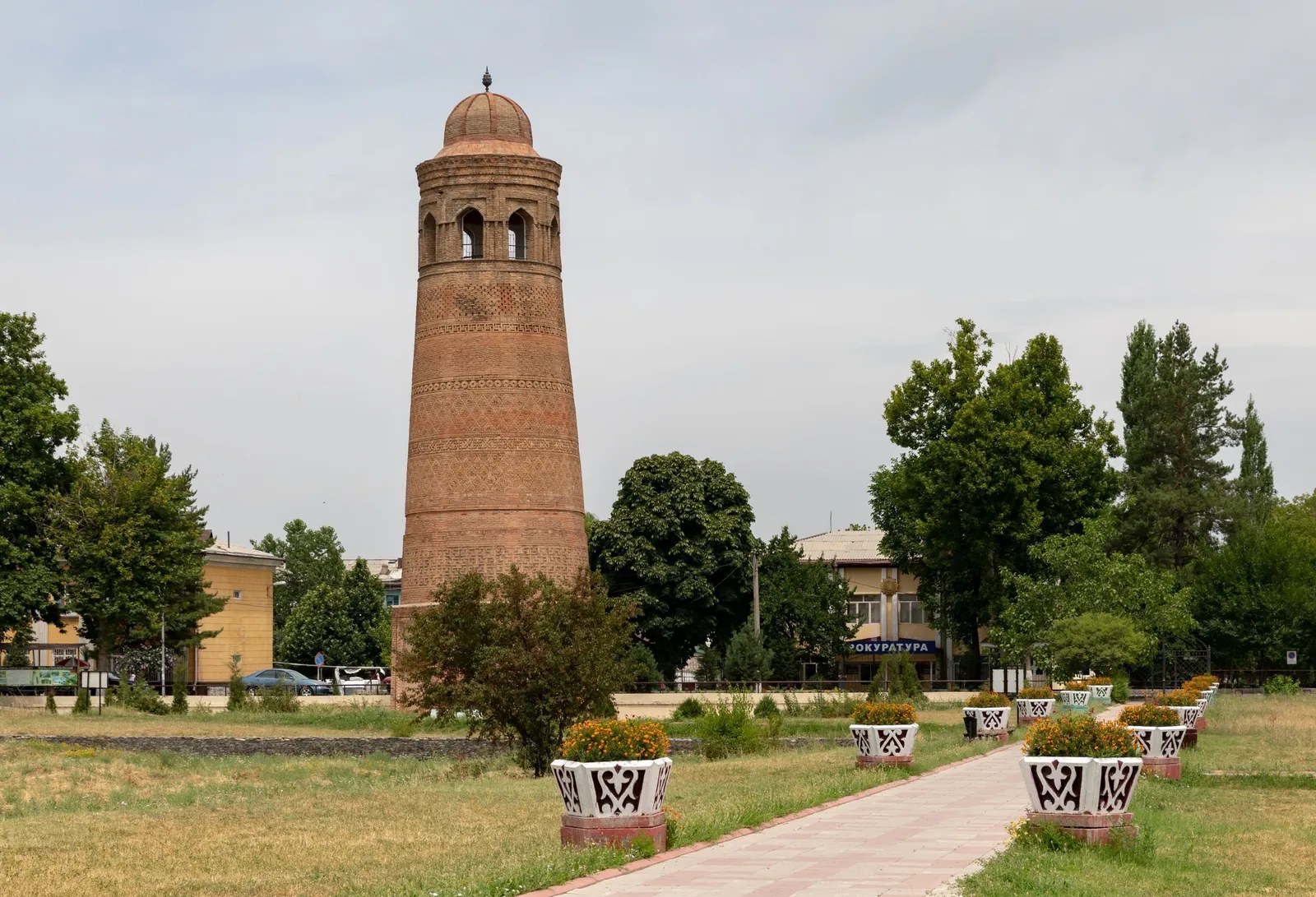 Ein Backsteinminarett mit Ziermauerwerk und rundem Abschluss über Rasen und Blumenbeeten.