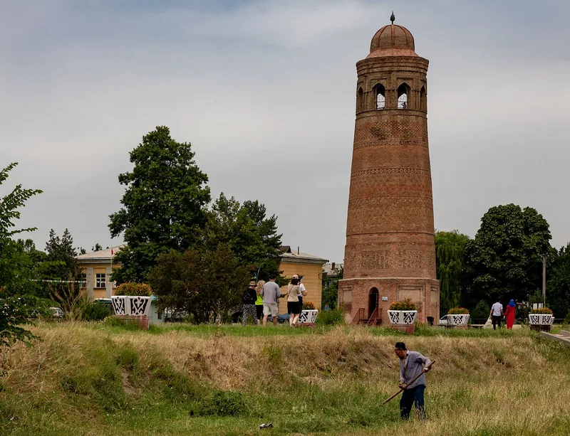 Minaret en brique sur une cour verte avec des bouquetières ; les visiteurs se tiennent le long du chemin, un homme travaille en dessous.