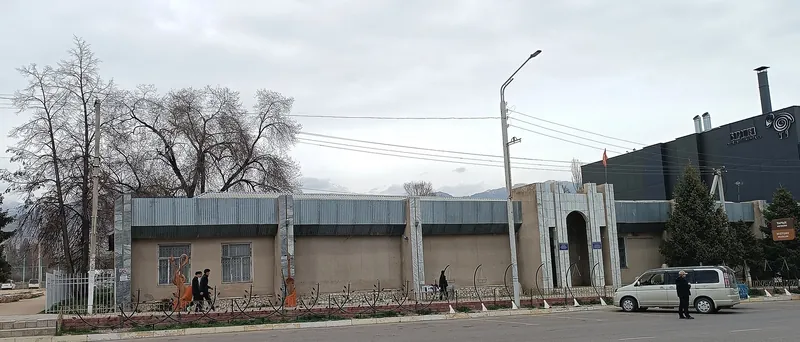 Невысокое музейное здание у городской улицы, слева деревья без листвы.