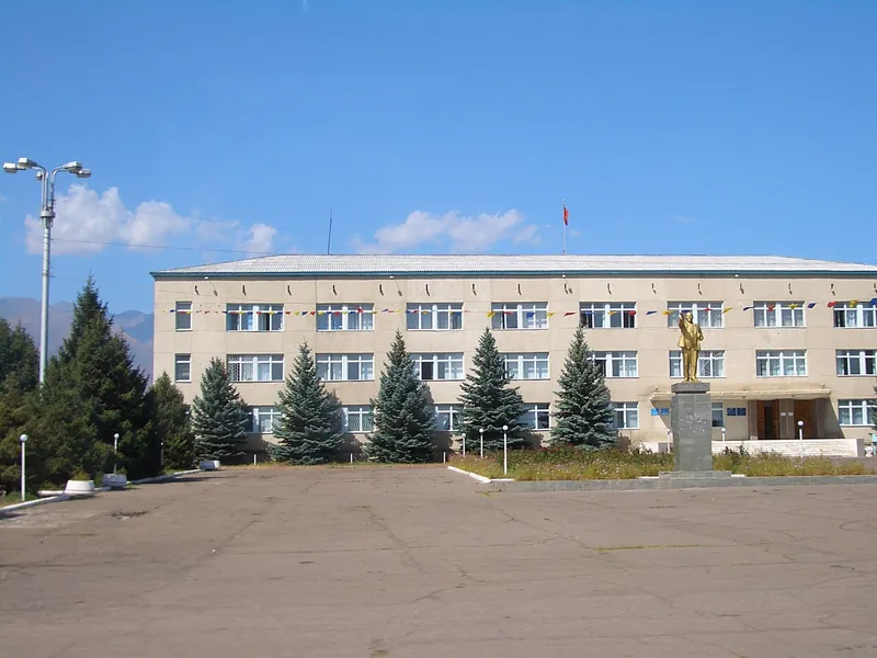 Трёхэтажное светлое здание за площадью, перед фасадом невысокие ели и флаг.