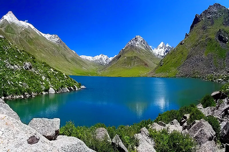 Il Lago Turchese Lago Besh-Tash è circondato da verdi pendii e picchi rocciosi; Ci sono grandi pietre sulla riva vicina.
