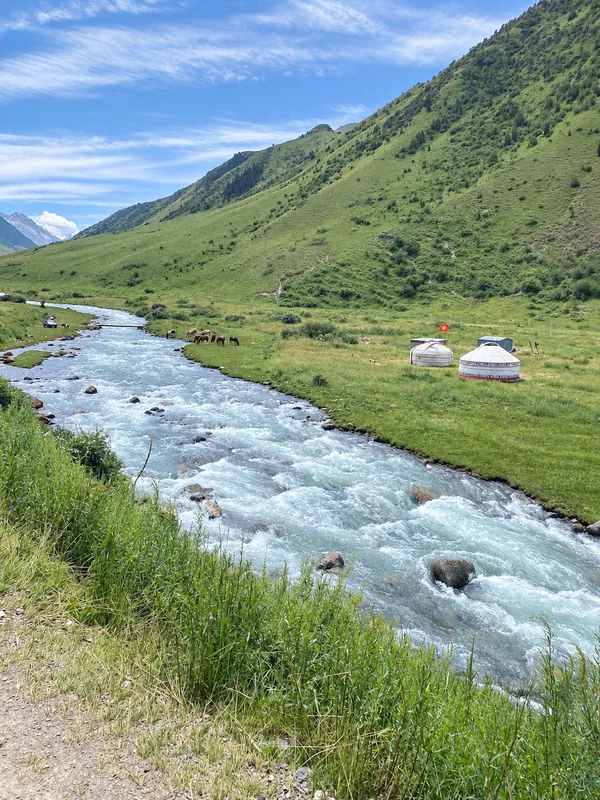 Fiume in una valle di montagna con yurte sulla riva, sfumature verdi primaverili dei pendii.