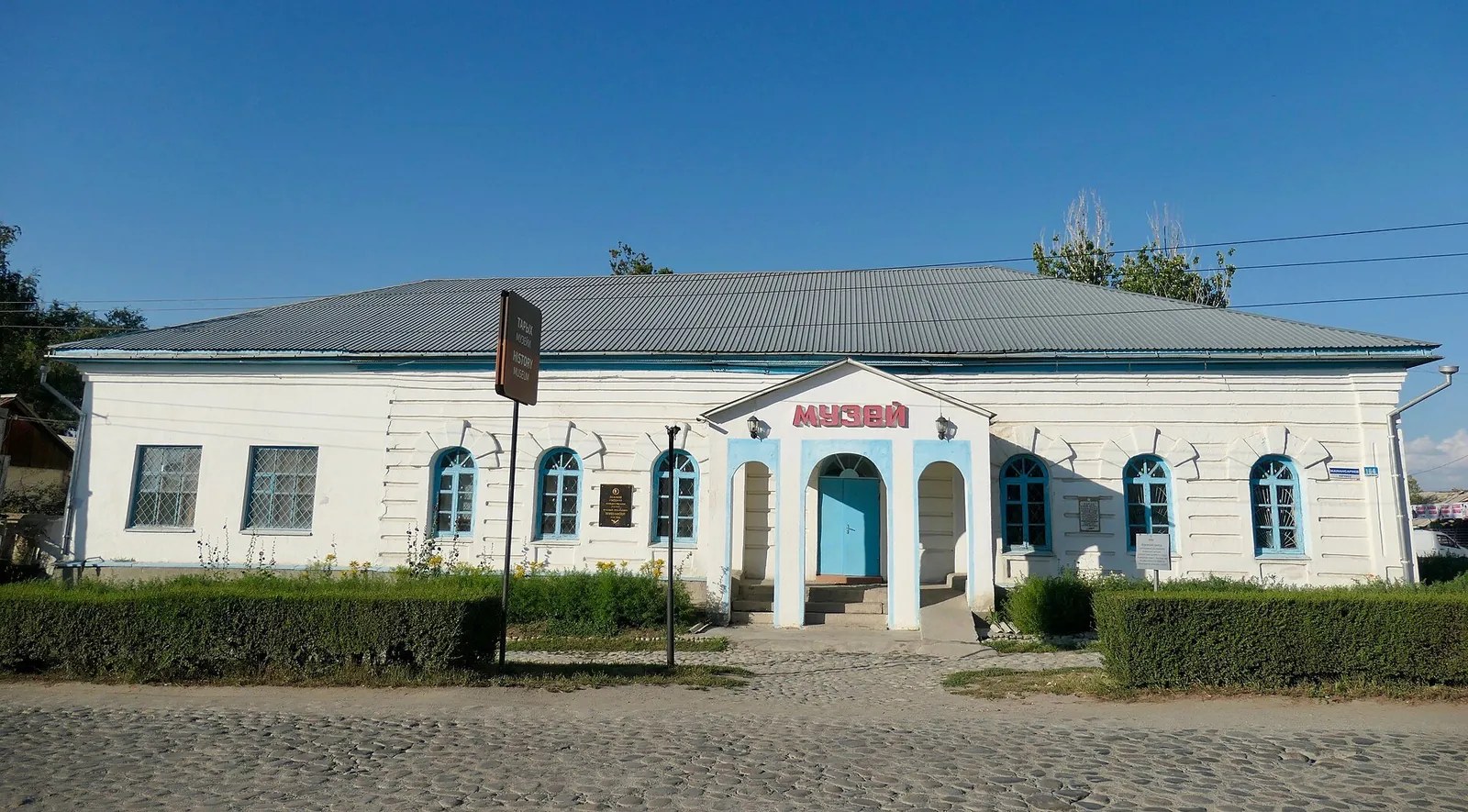 Museo blanco con ventanas y entrada azules y arbustos recortados ante la fachada.