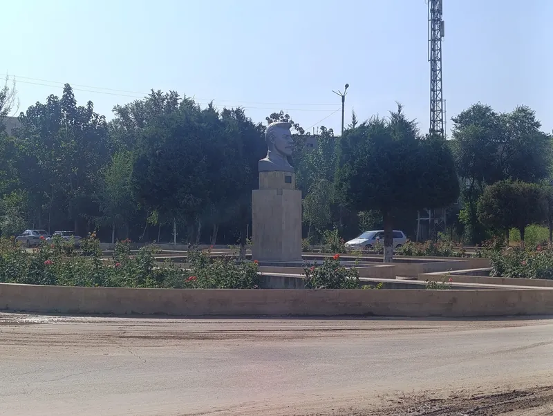 Busto sobre un alto pedestal entre rosas y árboles verdes junto a una calle urbana.
