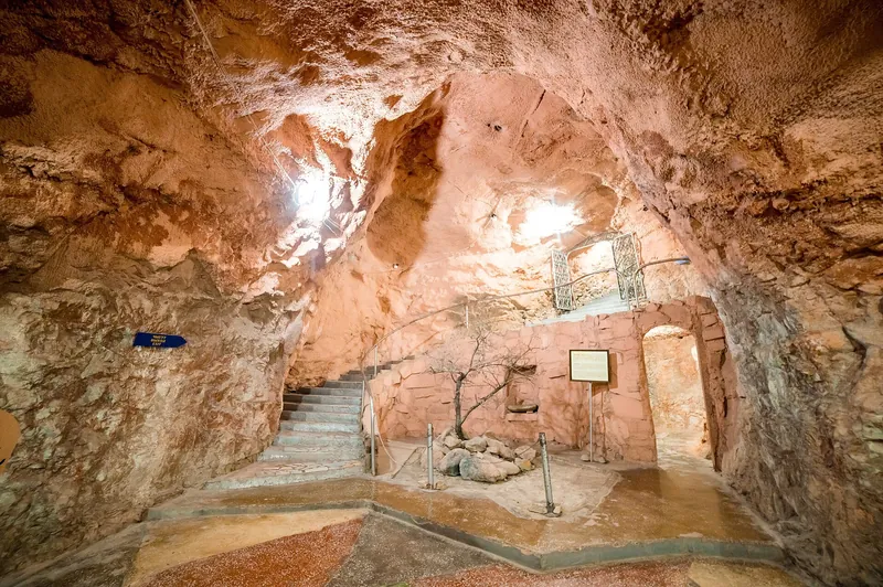 Una escalera se eleva entre las paredes irregulares de la cueva y una puerta de madera se encuentra junto a la vitrina iluminada.