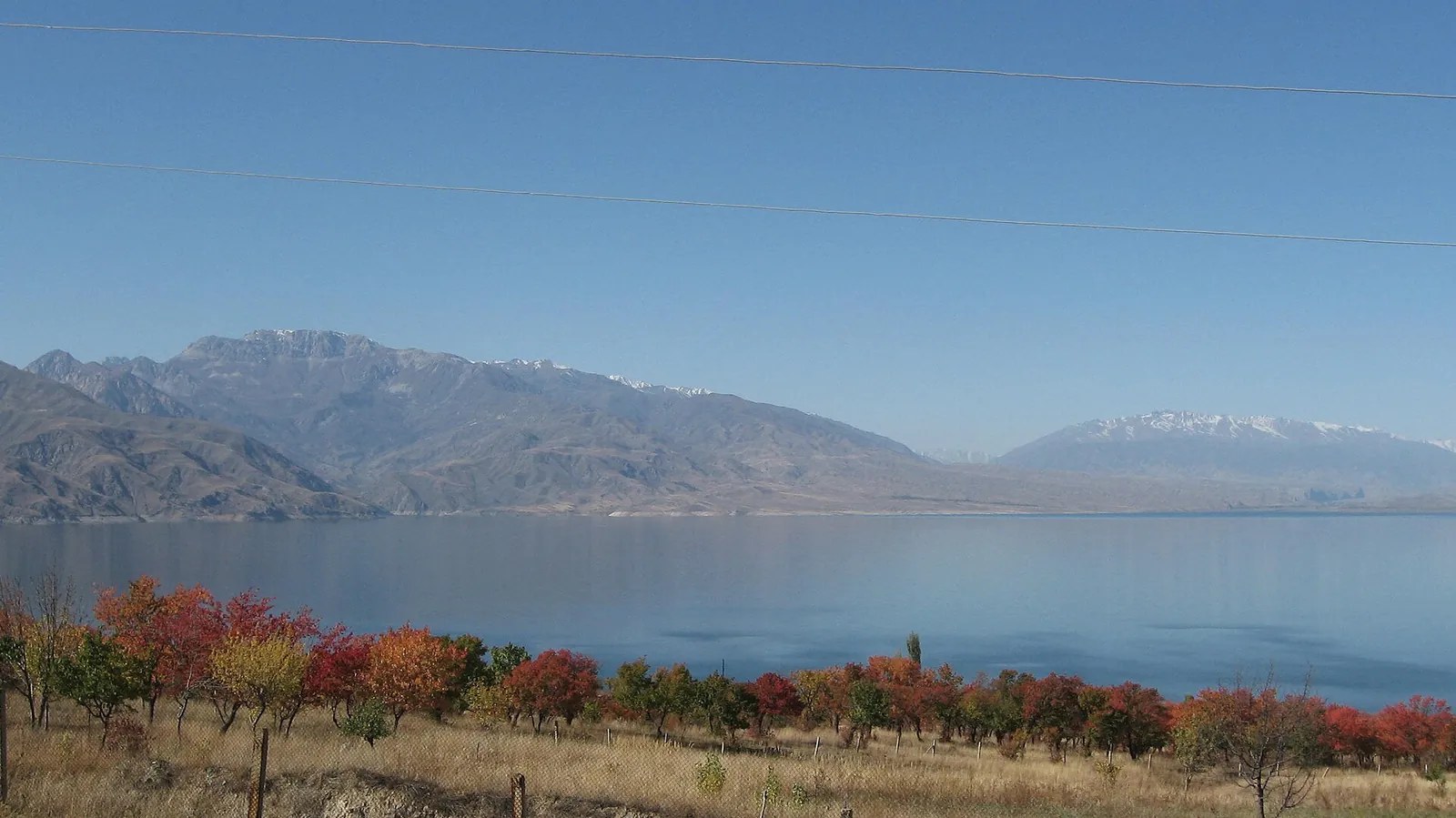 Herbstliche Bäume stehen über dem ruhigen Stausee, hinter dem fernen Ufer liegen Berge.