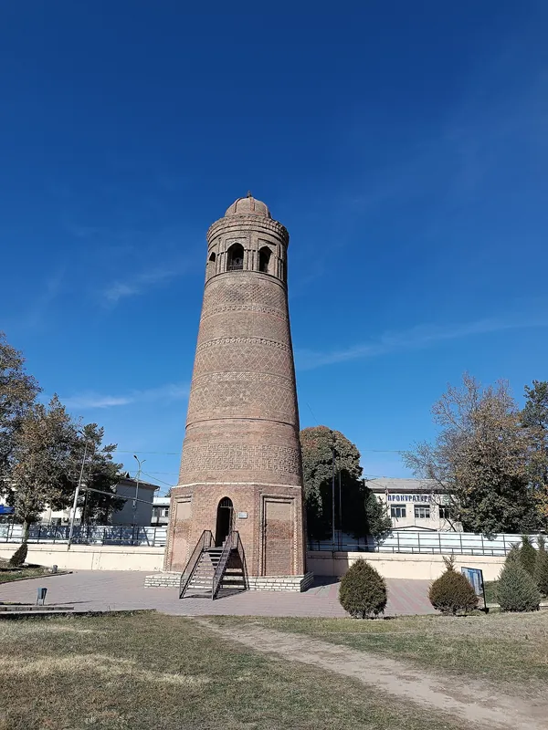 Minareto in mattoni di Uzgen contro il cielo blu, con gradini e recinzione chiara alla base.