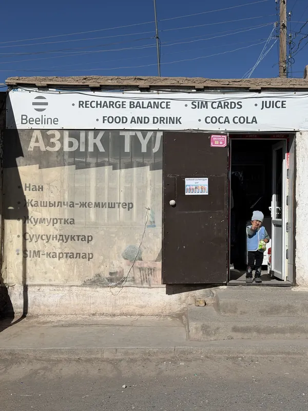 Un piccolo negozio a Sary-Mogol con un cartello sopra l'ingresso, una porta aperta e cavi sopra il tetto.