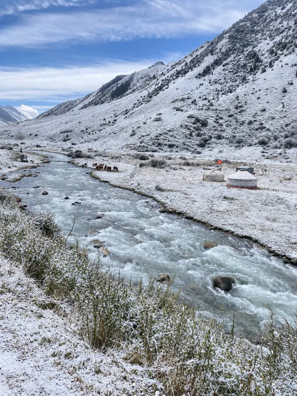 Fluss in einem Gebirgstal mit Jurten am Ufer, leichtem Schnee an den Hängen und Gras.