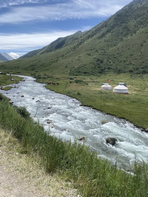 Ein weißer, reißender Fluss fließt durch ein grünes Tal; am anderen Ufer stehen zwei helle Jurten.